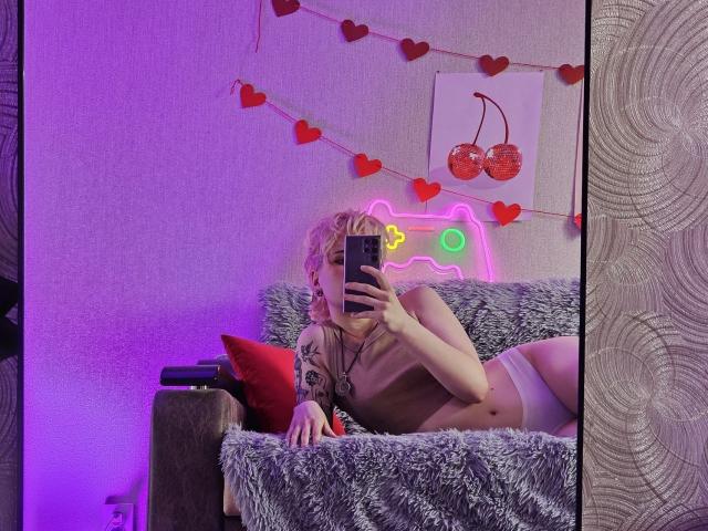 ShellyLove69 with blonde hair, lying on a gray fur-covered couch, taking a mirror selfie, wearing white panties, showing a tattoo on her right arm, purple and red lights.