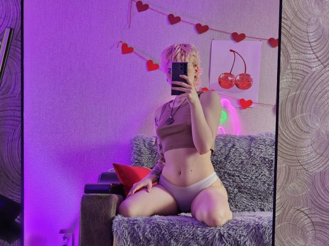 ShellyLove69, blonde with short hair, wears a tan sports bra and white panties, kneeling on a gray fuzzy sofa, taking a mirror selfie, showing her tits and pussy.