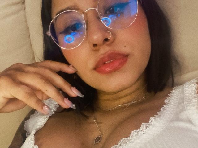 AshleyMayer with blue-eyed face filter, wearing round glasses, nose ring, and white lace top, showing off her shiny pink lips and cute cleavage, with a hand on her cheek.