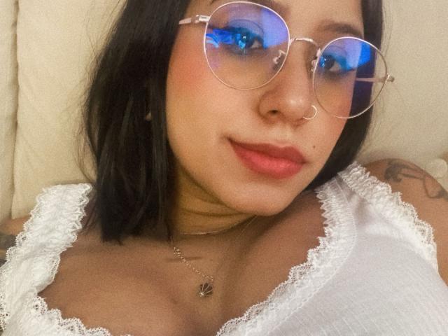 AshleyMayer with round glasses, blue-tinted eyes, and pink lipstick, wearing a white lace top, showing her tits, has black hair, a nose ring, and a tattoo.