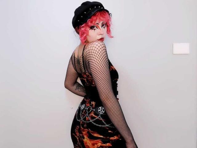 GothVioleth con i capelli ricci rosa, un cappello nero con borchie, maniche a rete, un vestito con motivo a fiamma, catene e un pentagramma sui glutei, che guarda indietro per spalla..