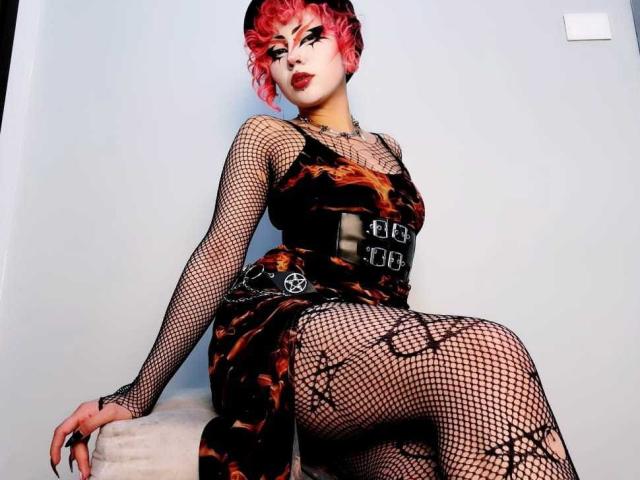 GothVioleth con cabello rizado rosa, vestido de malla negra y un vestido negro y naranja estampado, sentada en un taburete blanco, mostrando su trasero y sus pechos cubiertos de malla..