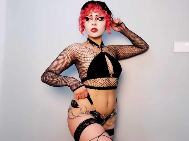 GothVioleth, avec ses cheveux roses bouclés, son haut en résille noire, son soutien-gorge noir, sa ceinture à chaîne, son bonnet noir et ses jarretières, montrant ses seins et ses fesses, debout devant un mur blanc..