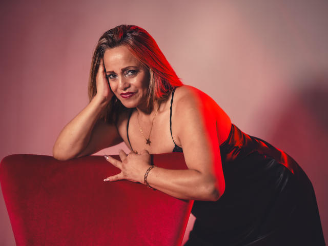 MaryMidori with red highlights, leaning on a red chair, wearing a black, low-cut dress, showing her tits, hand on chair, smiling, red and pink lighting, bracelet.