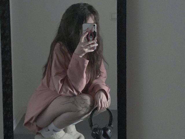 MissSpider with long brown hair, squatting, wearing a pink shirt, white socks, and showing a tattoo on her thigh, holding a phone, with headphones on the floor.