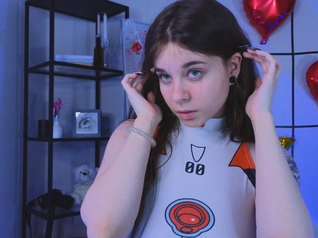 MissSpider with pale skin, blue eyes, and brown pigtails, wearing a white sleeveless top with a circular orange design, touching her ears, and looking slightly confused.