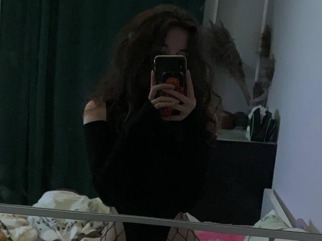Autoportrait de MissSpider avec des cheveux bruns bouclés, portant un haut noir à une épaule, tenant un téléphone, se cachant le visage, avec des bas en résille visibles. Pièce sombre, rideau vert..