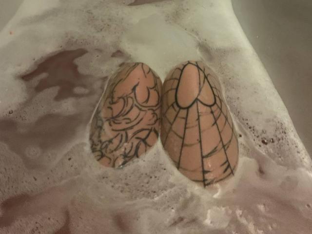 De tatoeages van Miss Spider in een badkuip, twee eieren met ingewikkelde zwarte lijntatoeages, gedeeltelijk ondergedompeld in zeepwater. Haar roze huid is zichtbaar door de bubbels..