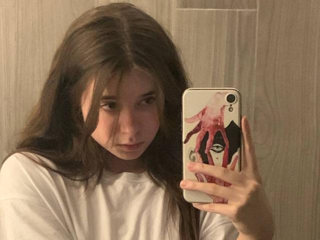 MissSpider with long brown hair, white shirt, holding phone with pink octopus case, looking in mirror, serious expression, wooden background. Phone has dual camera.
