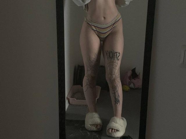 MissSpider's reflection in a mirror, wearing striped panties, white slippers, and showing tattooed thighs with "SWEET" on one leg and spider web designs on the other.