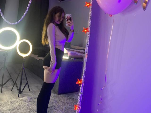 MissSpider with long brown hair, wearing a sheer top, black skirt, and thigh-high socks, taking a mirror selfie, showing her ass, in a room with ring lights.