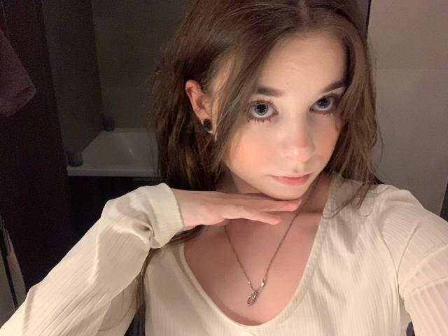 MissSpider with brown hair, blue eyes, and black ear gauges, wearing a white ribbed shirt, touching her chin, necklace with small pendant, soft lighting, slight smile, bathroom setting.