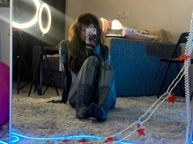 MissSpider with long brown hair, sitting on a carpet, wearing a gray top and black boots, taking a selfie with a ring light and blue LED strip in the mirror.