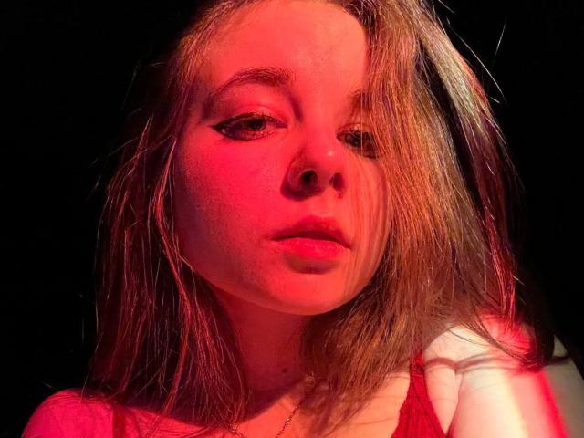 MissSpider has wet, dirty blonde hair, red-lit face, and serious expression. She's wearing a red top, with subtle makeup and a gold necklace, against a dark background.