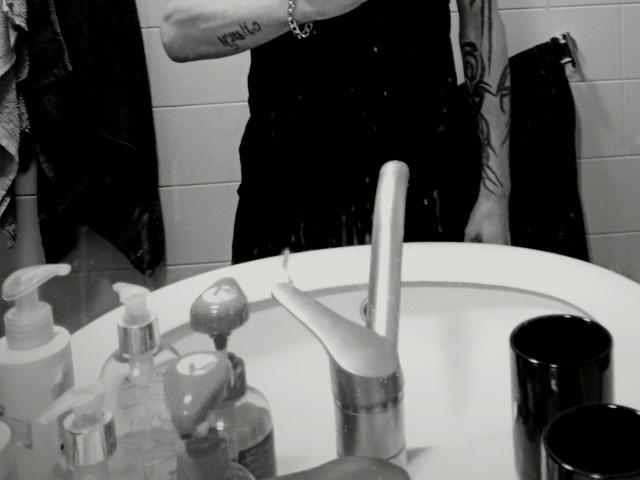 El brazo de MilfAnaLyzer-hot tiene un tatuaje que dice "wolo", lleva una camiseta negra sin mangas, pantalones negros mojados, y está de pie junto a un lavabo con un grifo, un cepillo de dientes y varios artículos de tocador..