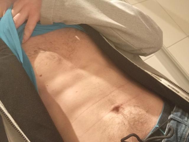 MilfAnaLyzer-hot, con su abdomen plano, cubierto de vello, tonificado y con un pequeño ombligo, vistiendo una camisa azul y una chaqueta gris con cremallera, ligeramente levantada para mostrar su torso sin pechos, cubierto de vello..