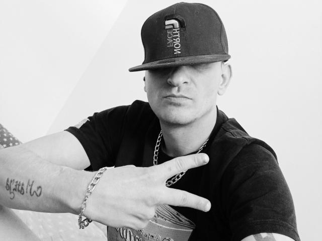 Fotografía en blanco y negro de MilfAnaLyzer, con un gorro de béisbol oscuro, un collar de cadena y un tatuaje en su brazo derecho, haciendo un gesto con la mano al estilo rock and roll. Su rostro es serio..