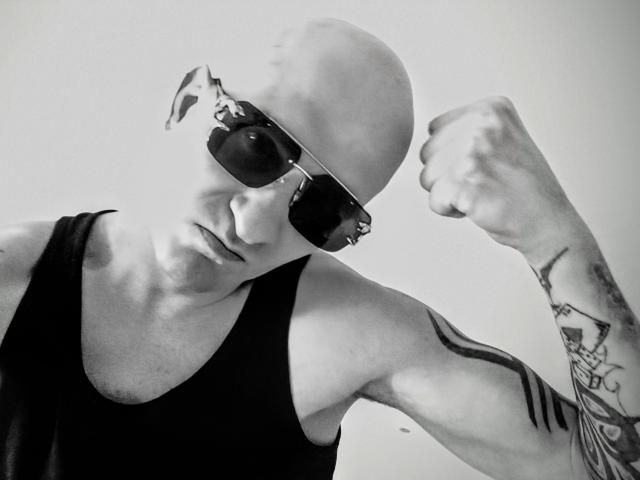 Fotografía en blanco y negro de MilfAnaLyzer, con la cabeza calva, usando gafas de sol oscuras, una camiseta negra sin mangas y mostrando un brazo tatuado mientras flexiona, con una expresión seria y ligeramente molesta..