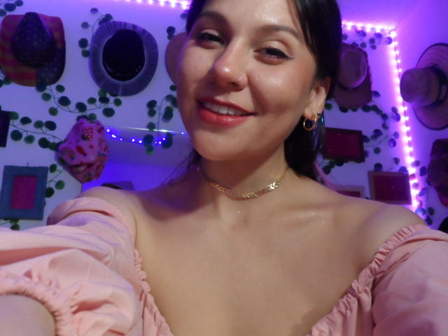SugarMuse69 with shiny skin, pink off-shoulder top, small gold hoop earrings, nose ring, and subtle smile, wearing a gold chain necklace, under purple LED lights.