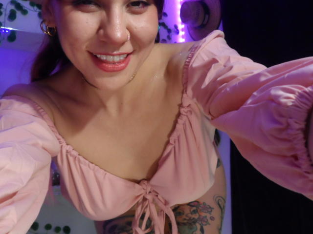 SugarMuse69 with a cute smile, pink off-shoulder top, small tits, tattoo on right arm, nose ring, purple lighting, and a slightly sweaty face. She's leaning forward.