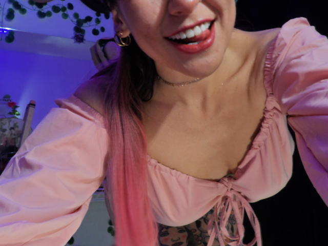 SugarMuse69 with pink-tipped hair, smiling with tongue out, wearing a pink off-shoulder top, showing her boobs, choker, and gold hoop earrings in a dimly lit room.