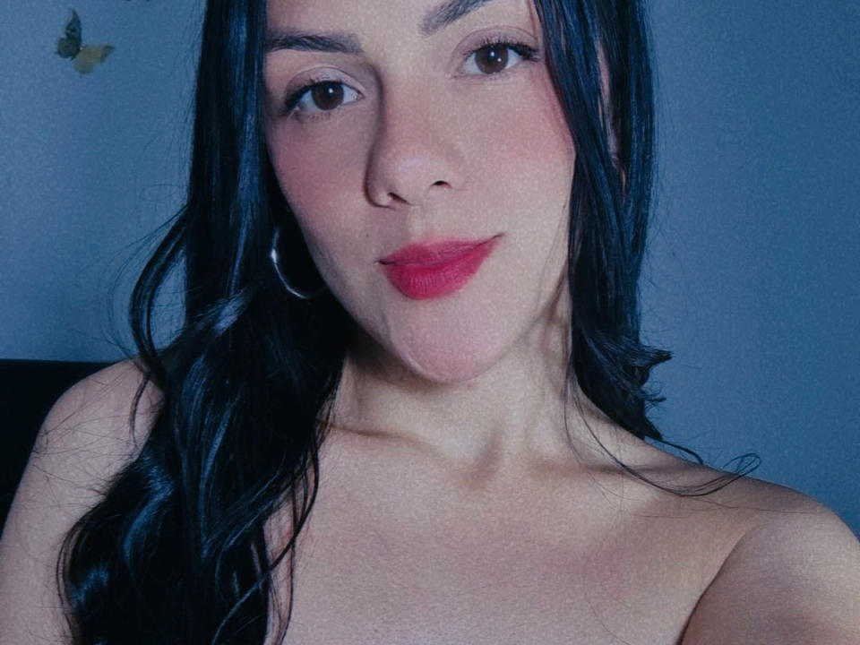 LulyLiz has fair skin, long black hair, and red lipstick. She's looking at the camera with a slight smile, wearing a silver hoop earring, and showing her bare shoulders.