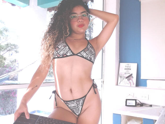 LizzaTaila with curly brown hair, glasses, and black patterned bikini, showing off her small tits and slim pussy. She's standing in a sunlit room, hand on head.