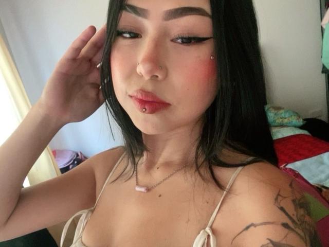 BriianaBx hat schwarzes Haar, roten Lippenstift, Eyeliner, einen Nasenring und eine Lippenpiercing. Sie trägt ein weißes Oberteil, das Tattoos an ihrem rechten Arm zeigt. Ihre Hand berührt ihren Kopf..