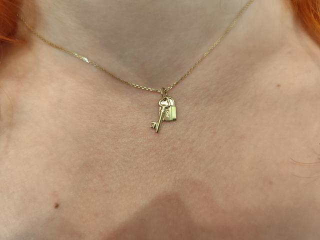 MoonBlossom's neck with a gold chain and a single key pendant, showing pale skin and red hair. Boobs in the background, slightly visible.