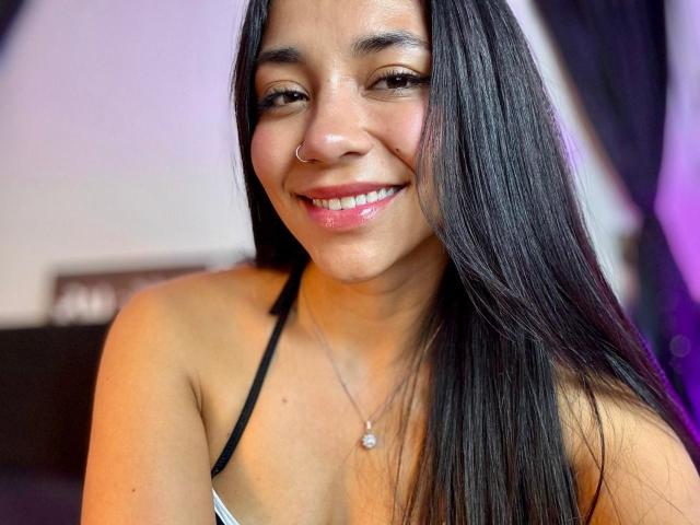 SophieDenisee tem cabelos pretos longos, um piercing no nariz e um colar de prata com uma pequena pérola. Ela está sorrindo, usando batom rosa, e tem um rosto bonito e atraente..