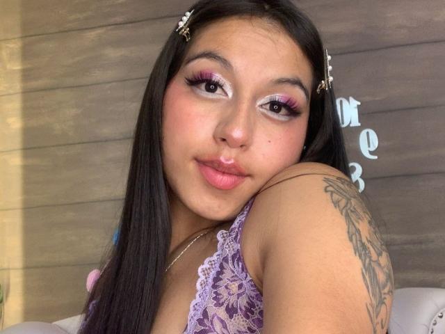 MadyJackson with long black hair, pink eyeliner, and purple lace top, has a tattoo on her right arm and a playful expression, showing her pouty pink lips.
