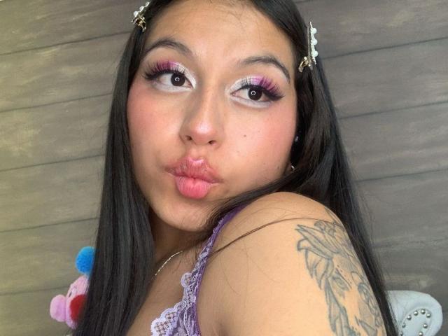 MadyJackson has pink and purple eyeliner, long black hair with flower hairpins, a tattoo on her right shoulder, and puffed pink lips. She's wearing a lace purple top.
