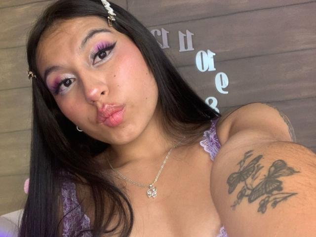 MadyJackson has pink eyeshadow, long black hair, and a tattoo on her arm with butterfly designs. She's pouting her lips and wearing a purple lace top.