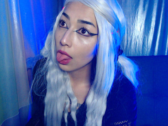 AlicePhantom, avec ses cheveux blancs, sa langue tirée et son eye-liner audacieux, a l'air mignonne et espiègle, vêtue d'un haut noir, dans un décor éclairé en bleu. Ses seins ne sont pas visibles..