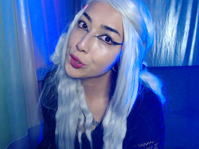 AlicePhantom with long white hair, thick black eyeliner, and pink lipstick, giving a cute pouty smile, wearing a black top, under blue and purple lighting.