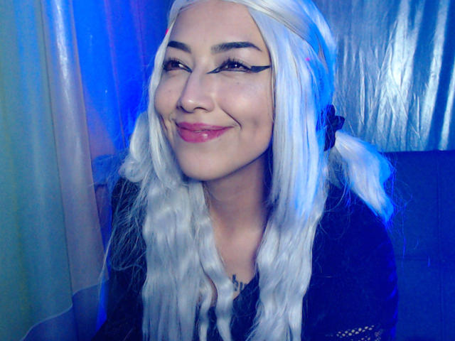 AlicePhantom with white hair, black eyeliner wings, and pink lipstick, smiling with red lips, wearing a black top, in a blue-lit setting. Her tits are small and she's cute.