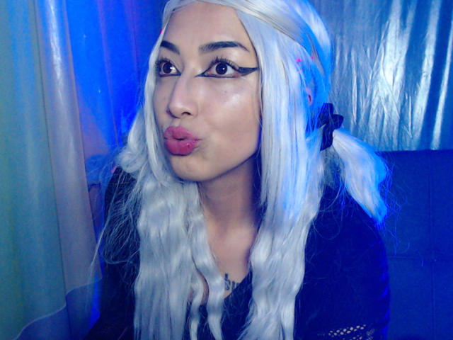 AlicePhantom with white hair in pigtails, black eyeliner, and pink lips, pouting her mouth. She's wearing a black top, and the blue lighting highlights her face.