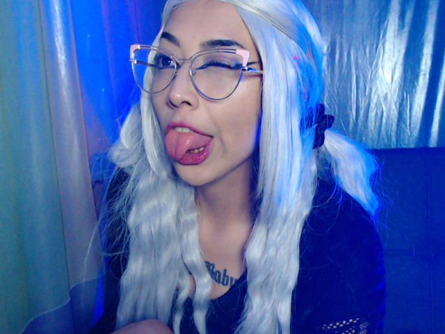 AlicePhantom met lang wit haar, een bril, haar tong uit, en een zwarte top, waarbij een tongpiercing zichtbaar is, in een selfie met blauw licht. Haar borsten zijn gedeeltelijk zichtbaar..