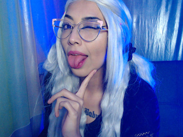 AlicePhantom with white hair, glasses, and tongue out, touching her chin, showing a tattoo on her chest. She's wearing a black top, looking cute and playful.