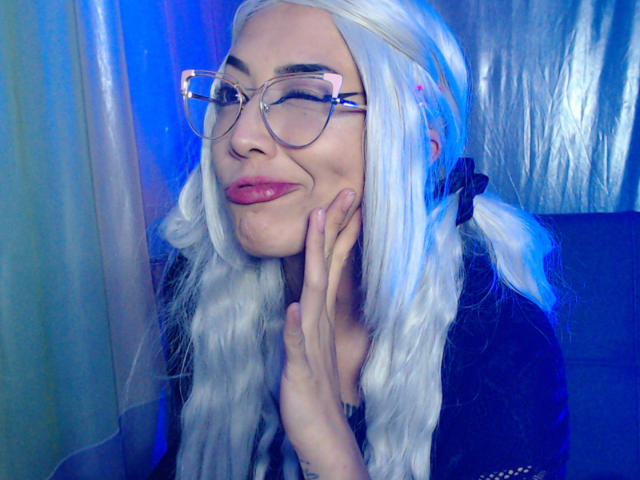 AlicePhantom met wit, golvend haar, roze lippenstift en een bril met heldere glazen, die haar wang aanraakt met haar hand en lichtjes glimlacht. Ze draagt een zwarte top, en op de achtergrond is blauw licht te zien..