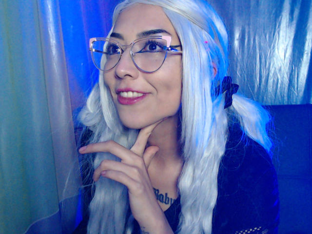 AlicePhantom, avec de longs cheveux blancs, des lunettes claires, un sourire, du rouge à lèvres rose, un haut noir, les mains près du menton, un éclairage bleu et violet, qui a l'air mignonne et séduisante..