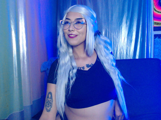 AlicePhantom met lang wit haar, een bril, een zwart topje dat haar borsten laat zien, een tatoeage op haar linkerarm, lachend, blauw licht, roze lippenstift, zittend tegen donkere gordijnen..