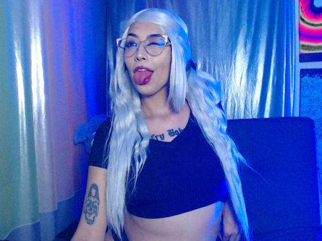 AlicePhantom with long white hair, glasses, black crop top, tongue out, tattoo on right arm, blue lighting, and a cute, playful expression. Boobs visible under top.