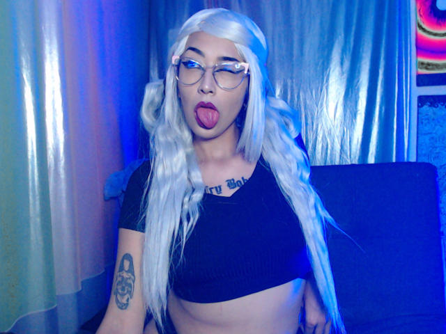 AlicePhantom met lang, golvend wit haar, een bril, een zwart topje, haar tong uitgestoken, een tatoeage op haar rechterarm, en blauwe belichting, waarbij kleine borsten en een platte achterkant te zien zijn..