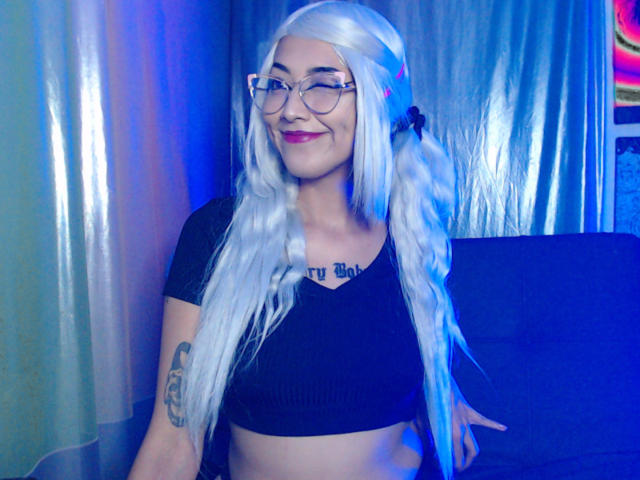 AlicePhantom met lange witte vlechtjes, een bril, een zwart topje, lachend, met kleine borsten, een tatoeage op de linkerarm, blauwe en roze verlichting, zittend in een kamer..