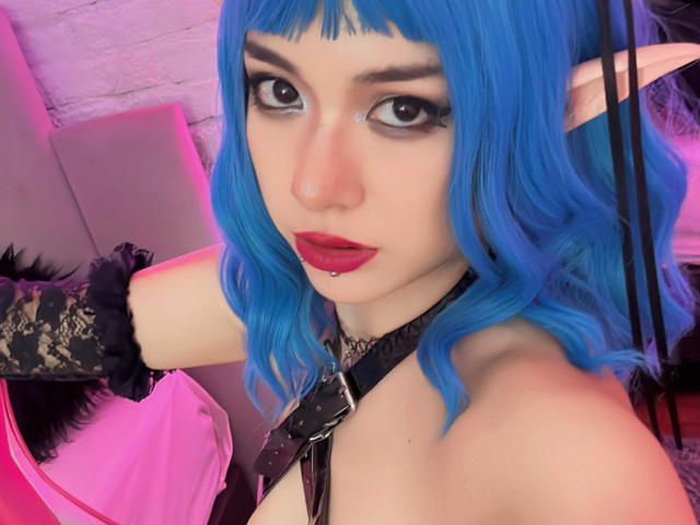 SabriPrettty with blue hair, elf ears, black lace gloves, red lipstick, and a black harness, looking up with a seductive expression, showing her bare shoulders and chest.
