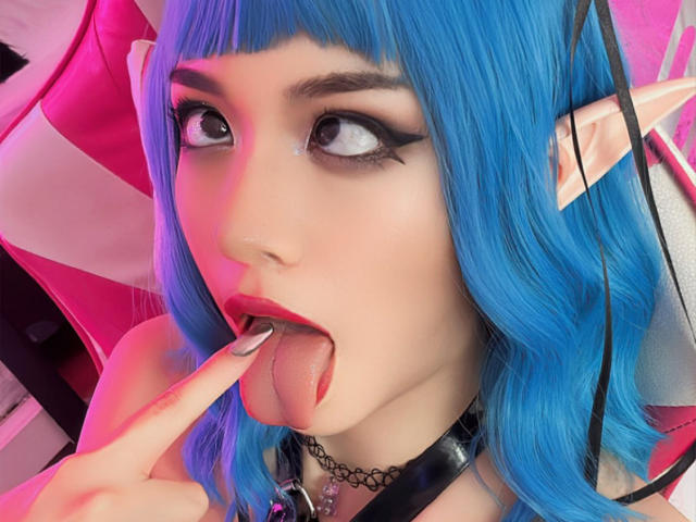 SabriPrettty with blue hair, thick black eyeliner, and pink highlights, tongue out, licking finger, wearing a black choker, and pink-tinged pink chair in the background.
