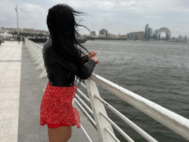 AmelieMuller with long black hair, wearing a black leather jacket and red floral skirt, leans on a white railing by the water, her ass slightly visible.