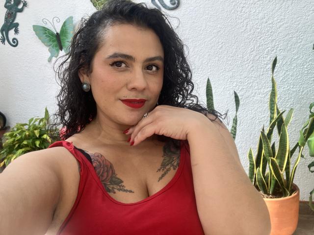 Tamarajoones with curly black hair, red lipstick, and red tank top showing tattoos on her tits, resting chin on hand, smiling at camera, surrounded by potted plants.
