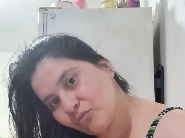 CressidaX has wet black hair, brown eyes, and a slightly pouty face with shiny skin, wearing a green patterned top, standing in front of a white fridge.